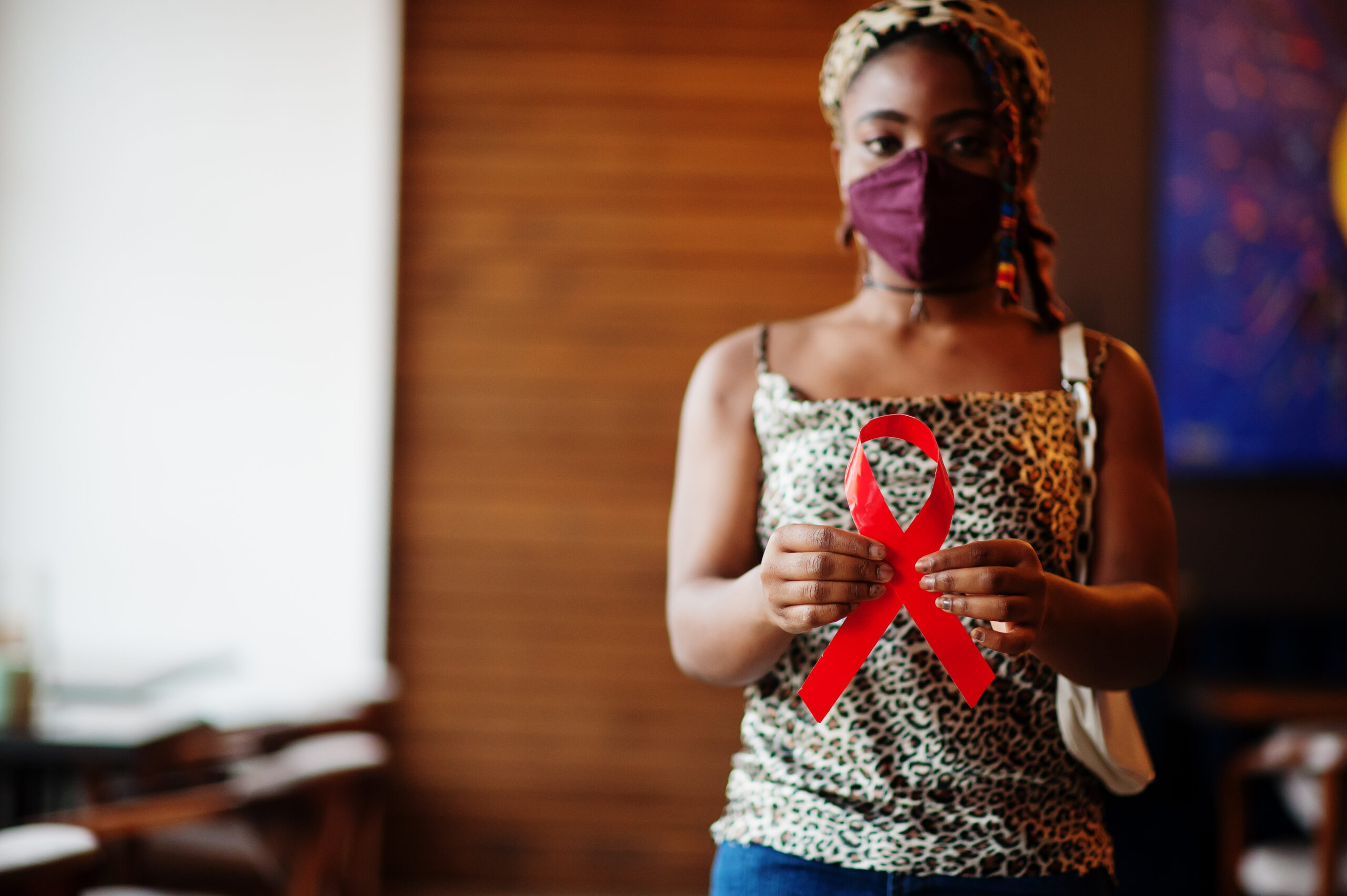 AIDS awareness. Stylish african american woman with dreadlocks afro hair, wear face protect mask and headdress leopard scarf, hold red ribbon.